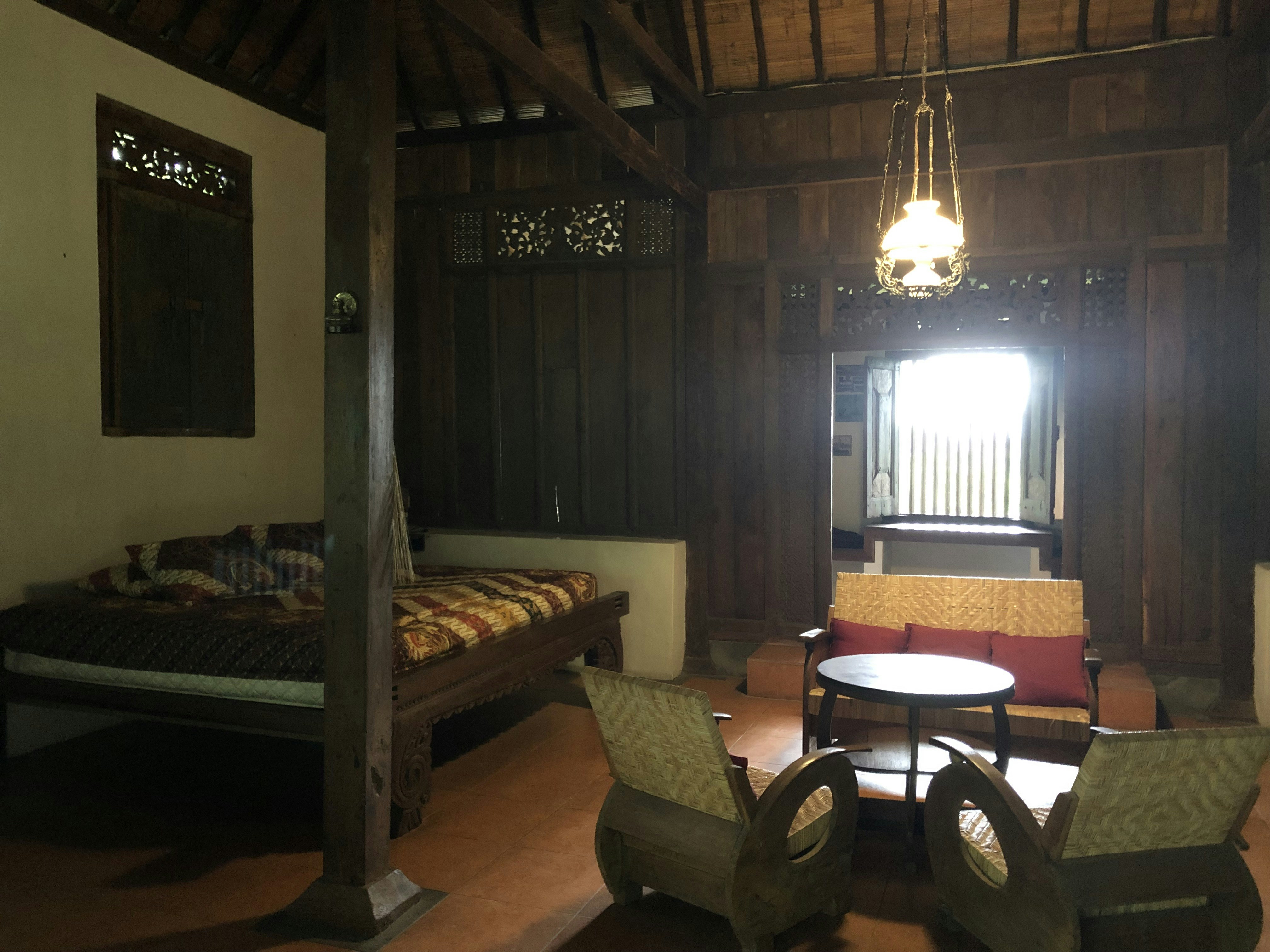 A cozy, dimly lit room with wooden walls and ceiling featuring traditional design elements. There is a bed on the left side with a patterned quilt, and in the center, a seating area consisting of two armchairs and a round table facing a loveseat with red pillows. A decorative light fixture hangs from the ceiling, casting a warm glow.
