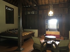 A cozy, dimly lit room with wooden walls and ceiling featuring traditional design elements. There is a bed on the left side with a patterned quilt, and in the center, a seating area consisting of two armchairs and a round table facing a loveseat with red pillows. A decorative light fixture hangs from the ceiling, casting a warm glow.