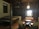A cozy, dimly lit room with wooden walls and ceiling featuring traditional design elements. There is a bed on the left side with a patterned quilt, and in the center, a seating area consisting of two armchairs and a round table facing a loveseat with red pillows. A decorative light fixture hangs from the ceiling, casting a warm glow.