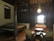 A cozy, dimly lit room with wooden walls and ceiling featuring traditional design elements. There is a bed on the left side with a patterned quilt, and in the center, a seating area consisting of two armchairs and a round table facing a loveseat with red pillows. A decorative light fixture hangs from the ceiling, casting a warm glow.