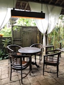 An outdoor seating area with a round wooden table surrounded by four wooden chairs. White curtains are drawn open, revealing a lush garden with bamboo plants. A hanging light fixture is suspended above the table, and the floor is made of stone tiles. A wooden door and potted plants add to the rustic ambiance.