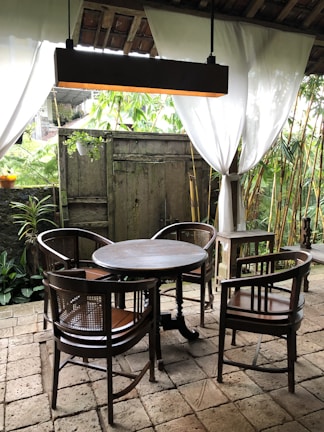 An outdoor seating area with a round wooden table surrounded by four wooden chairs. White curtains are drawn open, revealing a lush garden with bamboo plants. A hanging light fixture is suspended above the table, and the floor is made of stone tiles. A wooden door and potted plants add to the rustic ambiance.