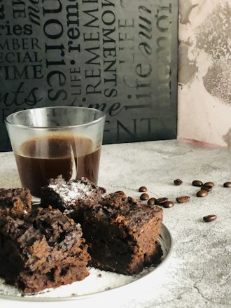 A wooden surface holds a glass of dark coffee and a plate of chocolate brownies dusted with powdered sugar. Scattered coffee beans add a rustic touch. The dark-themed background features various typography.