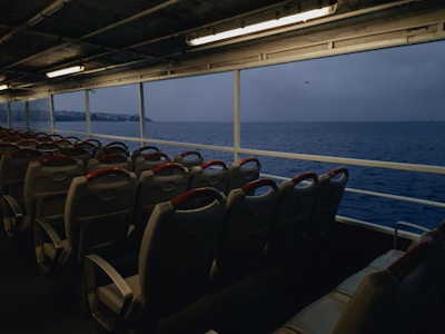 An empty ferry interior with rows of seats facing large windows, offering a view of the open sea. The sky appears overcast, creating a moody atmosphere. The interior is dimly lit with fluorescent lights overhead, and the seats have red headrests.