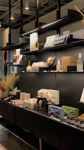 A modern retail display features various packaged goods arranged neatly on black shelves. There are bottles, wrapped boxes, and jars, along with decorative elements like pampas grass. The lighting highlights the products, adding a sophisticated and minimalistic vibe.