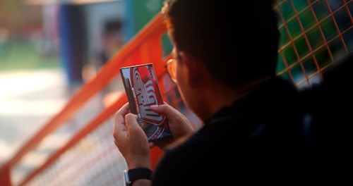 A person holding and interacting with a mobile device, with a focus on the screen displaying colorful graphics. The background contains a blurred view of an outdoor setting with a grid-like orange structure.