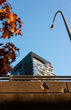 Modern architectural building with a distinct geometric design stands against a clear blue sky. A streetlamp arches over the scene, adding an urban element. In the foreground, the edge of a brown concrete structure can be seen, along with branches of a tree displaying autumnal foliage in shades of orange and red.
