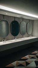 A public restroom features a row of five oval mirrors aligned above a long countertop with five sinks. The wall is covered with small, square, gray tiles. Soft lighting from the ceiling creates a calm ambiance. Geometric floor tiles add a modern touch to the overall design.