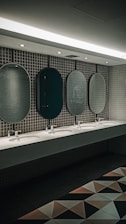 A public restroom features a row of five oval mirrors aligned above a long countertop with five sinks. The wall is covered with small, square, gray tiles. Soft lighting from the ceiling creates a calm ambiance. Geometric floor tiles add a modern touch to the overall design.