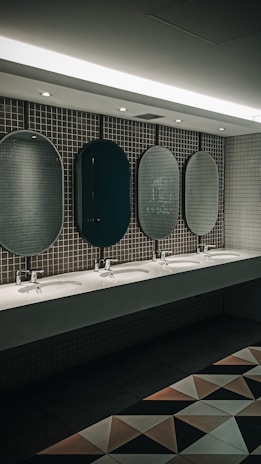 A public restroom features a row of five oval mirrors aligned above a long countertop with five sinks. The wall is covered with small, square, gray tiles. Soft lighting from the ceiling creates a calm ambiance. Geometric floor tiles add a modern touch to the overall design.