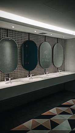 A public restroom features a row of five oval mirrors aligned above a long countertop with five sinks. The wall is covered with small, square, gray tiles. Soft lighting from the ceiling creates a calm ambiance. Geometric floor tiles add a modern touch to the overall design.