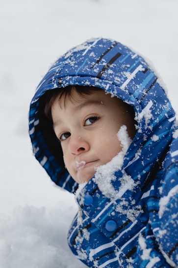 A child wearing a blue patterned winter coat looks towards the camera. The coat is covered with snow, and snowflakes are visible on the child's hair and face. The background is filled with more snow, suggesting a snowy outdoor setting.