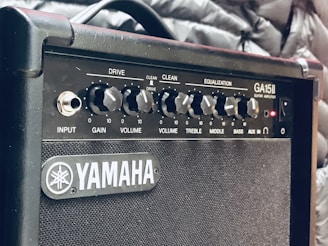 A close-up view of a Yamaha GA15II guitar amplifier with various control knobs labeled Drive, Gain, Volume, Treble, Middle, and Bass. The amplifier has a textured grille and a sleek black design. Surrounding the amplifier are what appears to be a quilted jacket or padded fabric.