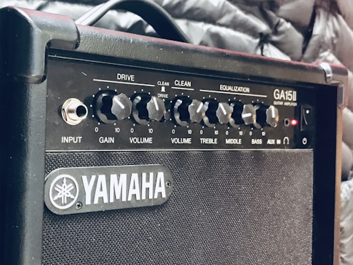 A close-up view of a Yamaha GA15II guitar amplifier with various control knobs labeled Drive, Gain, Volume, Treble, Middle, and Bass. The amplifier has a textured grille and a sleek black design. Surrounding the amplifier are what appears to be a quilted jacket or padded fabric.