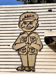 A stylized drawing of a smiling person wearing a uniform with a hat labeled 'BEST' is displayed on a textured, horizontal slatted surface. The figure has exaggerated facial features and is in a greeting posture with hands together.