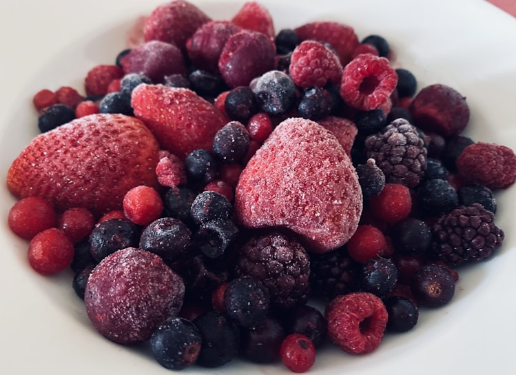 A close-up view of a variety of frozen berries, including strawberries, raspberries, blueberries, and blackberries, covered with a light layer of frost, placed on a white plate.