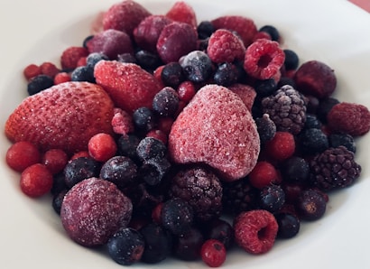 A close-up view of a variety of frozen berries, including strawberries, raspberries, blueberries, and blackberries, covered with a light layer of frost, placed on a white plate.