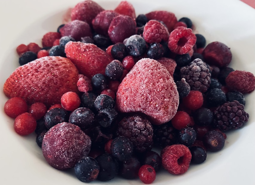 A close-up view of a variety of frozen berries, including strawberries, raspberries, blueberries, and blackberries, covered with a light layer of frost, placed on a white plate.
