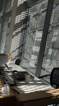 Sunlight casts patterned shadows through the perforated window panels, creating an intricate play of light and shadow on the interior surfaces. A desk is positioned against the windowsill, featuring various office essentials such as a laptop, desk phone, and phone charger. A disposable coffee cup and a bottle of sanitizer are also present on the desk. The overall setup suggests a modern office environment.