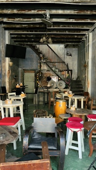 An empty, rustic cafe or bar interior features mismatched wooden tables and chairs, some with red or black cushions. A set of conga drums adds a musical element near a staircase, which leads up along a textured brick and plaster wall. Overhead, the ceiling exposes beams and piping, adding to the industrial aesthetic.