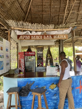 A rustic beach bar with a thatched roof and natural wooden decor. The bar, called 'Afro Beach Bar Mafia,' has stools, colorful ocean-themed wall art featuring fish and a starfish, and a variety of drinks in a cooler behind the counter. A person wearing a white tank top and shorts stands at the bar. The environment is tropical and relaxed.