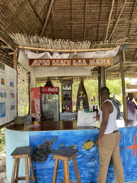 A rustic beach bar with a thatched roof and natural wooden decor. The bar, called 'Afro Beach Bar Mafia,' has stools, colorful ocean-themed wall art featuring fish and a starfish, and a variety of drinks in a cooler behind the counter. A person wearing a white tank top and shorts stands at the bar. The environment is tropical and relaxed.