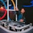 A DJ stands behind a DJ booth with turntables and mixers. He wears a jacket, headphones around his neck, and gestures energetically with one hand forward. The background features colorful circular designs on the wall with ambient lighting.
