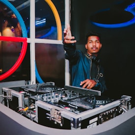 A DJ stands behind a DJ booth with turntables and mixers. He wears a jacket, headphones around his neck, and gestures energetically with one hand forward. The background features colorful circular designs on the wall with ambient lighting.