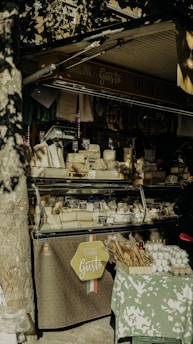 An outdoor market stall displays an array of cheeses and packaged goods. There is a variety of cheese blocks in a transparent display case, each labeled with small price tags. Packs of breadsticks and cartons of eggs are arranged on a table covered with a green and white patterned cloth. Dappled sunlight filters through the leaves of a tree, creating a mix of shadows and highlights. The stall's sign reads 'Origine di Gusto' and 'Salumi e Formaggi'.