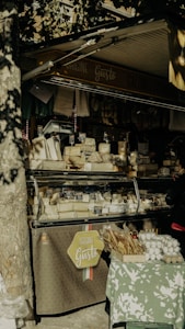 An outdoor market stall displays an array of cheeses and packaged goods. There is a variety of cheese blocks in a transparent display case, each labeled with small price tags. Packs of breadsticks and cartons of eggs are arranged on a table covered with a green and white patterned cloth. Dappled sunlight filters through the leaves of a tree, creating a mix of shadows and highlights. The stall's sign reads 'Origine di Gusto' and 'Salumi e Formaggi'.