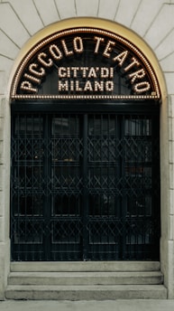 An arched entrance with ornate design features a sign that reads 'Piccolo Teatro Città di Milano,' illuminated with a series of lights. The doors are large with a vintage and intricate grid ironwork pattern, set into a stone facade.