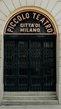 An arched entrance with ornate design features a sign that reads 'Piccolo Teatro Città di Milano,' illuminated with a series of lights. The doors are large with a vintage and intricate grid ironwork pattern, set into a stone facade.