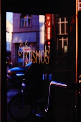 A dimly lit cafe entrance with reflections of a street scene visible in the glass. The word 'Kosmos' is prominently displayed in cursive writing on the glass. Through the reflection, a bicyclist and cars can be vaguely discerned on a street lined with classic architectural buildings.