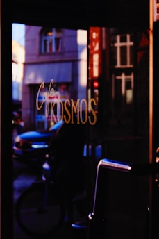 A dimly lit cafe entrance with reflections of a street scene visible in the glass. The word 'Kosmos' is prominently displayed in cursive writing on the glass. Through the reflection, a bicyclist and cars can be vaguely discerned on a street lined with classic architectural buildings.