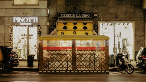A street scene at night featuring a pizzeria with a yellow awning labeled 'Pizzeria Da Gino' and a nearby fashion store with mannequins displayed. A covered outdoor dining area with fence-like sides is in front of the pizzeria. Several vehicles, including a motorcycle, are parked on the street alongside illuminated shop displays.