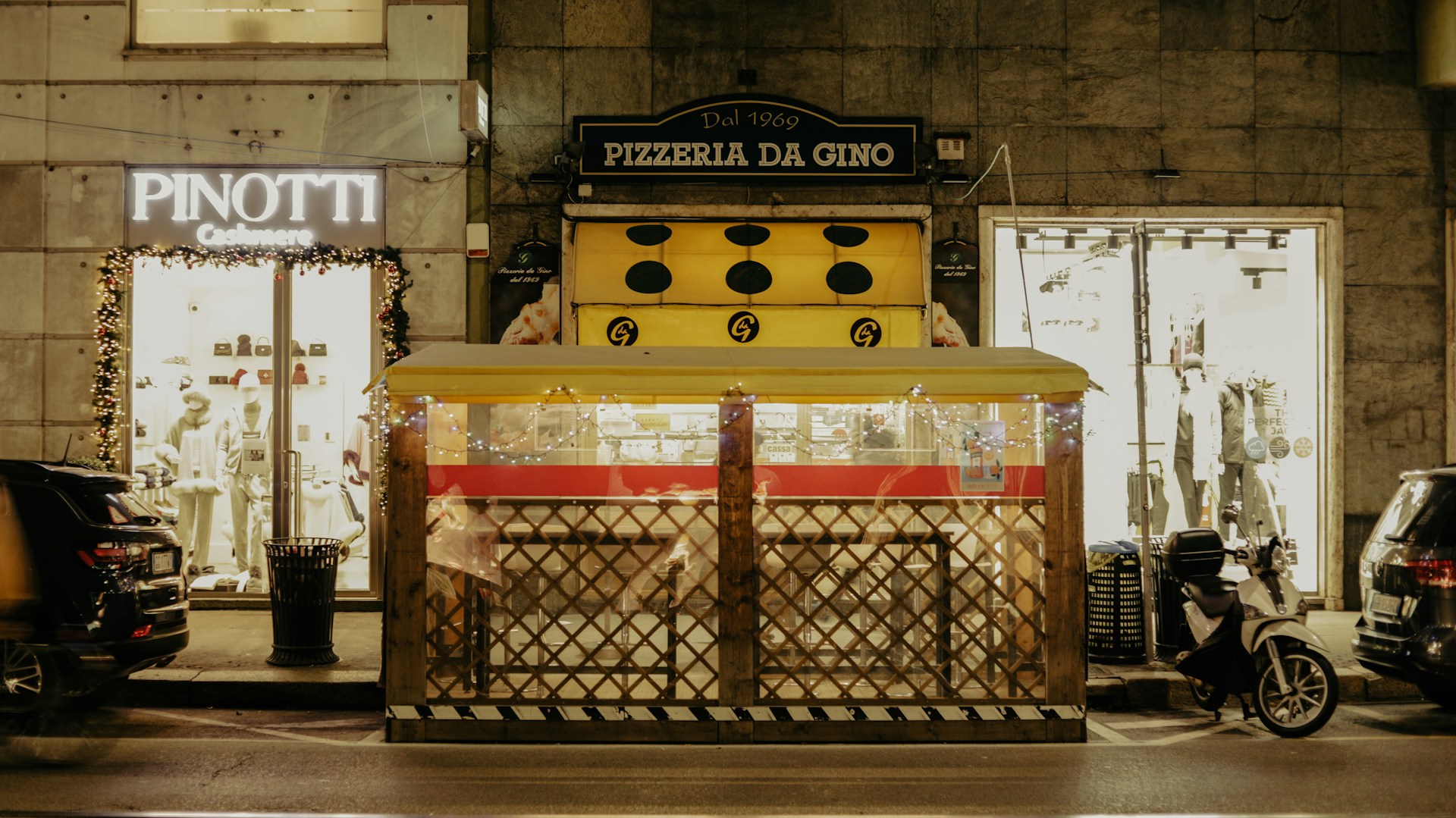 A street scene at night featuring a pizzeria with a yellow awning labeled 'Pizzeria Da Gino' and a nearby fashion store with mannequins displayed. A covered outdoor dining area with fence-like sides is in front of the pizzeria. Several vehicles, including a motorcycle, are parked on the street alongside illuminated shop displays.