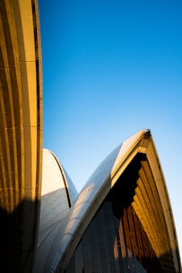 Architectural design with curved, overlapping structures set against a clear, blue sky. The building features a combination of smooth and textured surfaces, with distinct shadows cast by the sun.
