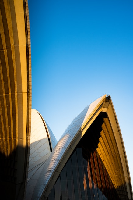 Architectural design with curved, overlapping structures set against a clear, blue sky. The building features a combination of smooth and textured surfaces, with distinct shadows cast by the sun.