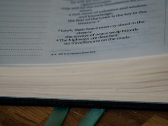 The close-up view of an open book reveals printed text with clear line spacing. The focus is on a portion of a page with highlighted text that reads, 'Look, their brave men cry aloud in the streets; the envoys of peace weep bitterly. The highways are deserted, no travelers are on the roads.' A ribbon bookmark is visible protruding from the bottom of the pages.
