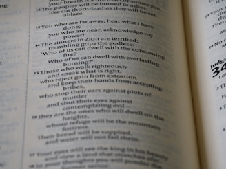 A close-up view of an open book displaying text in English. The text is organized in paragraphs and includes numbered verses, typical of a religious scripture. The paper is slightly off-white, and the typography is clear.
