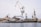 A shipyard with several large cranes and industrial structures is visible. The scene includes a vessel docked at the waterfront and multiple buildings with rusted surfaces. The sky is overcast, contributing to a muted tone.