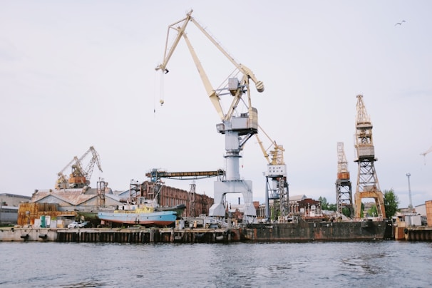 A shipyard with several large cranes and industrial structures is visible. The scene includes a vessel docked at the waterfront and multiple buildings with rusted surfaces. The sky is overcast, contributing to a muted tone.