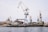 A shipyard with several large cranes and industrial structures is visible. The scene includes a vessel docked at the waterfront and multiple buildings with rusted surfaces. The sky is overcast, contributing to a muted tone.