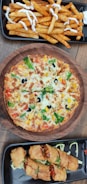 A wooden table showcases a round vegetable pizza topped with colorful vegetables like broccoli, bell peppers, tomato, and black olives, surrounded by golden cheese. Near the pizza, a platter of crispy seasoned fries drizzled with white sauce, and another platter with golden-brown fried spring rolls filled with a savory stuffing on a bed of shredded cabbage.