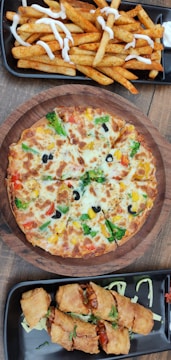 A wooden table showcases a round vegetable pizza topped with colorful vegetables like broccoli, bell peppers, tomato, and black olives, surrounded by golden cheese. Near the pizza, a platter of crispy seasoned fries drizzled with white sauce, and another platter with golden-brown fried spring rolls filled with a savory stuffing on a bed of shredded cabbage.