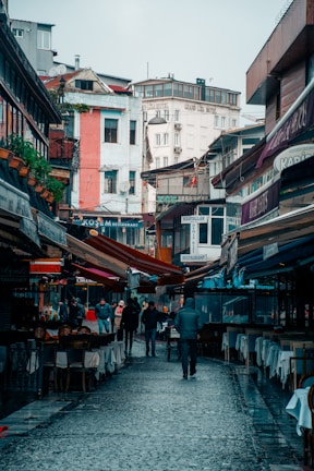 A bustling urban street lined with outdoor restaurants, with several people walking along the cobblestone path. The buildings in the background are multi-story with colorful facades, and awnings extend over the street, providing shade. Signs of various eateries are visible, creating a lively atmosphere.