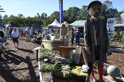 A lively outdoor event with various stalls and tents, surrounded by greenery and large trees. People are walking around, some in groups, enjoying the fair-like atmosphere. A life-sized pirate mannequin dressed in traditional pirate attire stands next to a display with gold coins, a globe, and a treasure chest. The ground is covered with wood chips, and small plants and flowers are integrated into the displays.