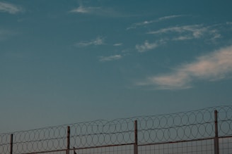 A barbed wire fence against a backdrop of a clear sky with scattered clouds. The fence consists of tall metal posts with circular barbed wire loops running along the top, suggesting a secure or restricted area.