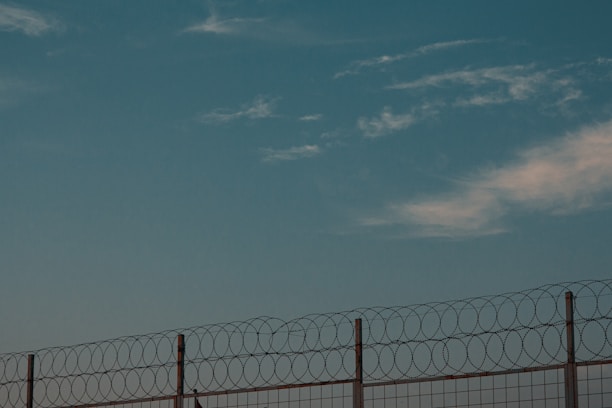 A barbed wire fence against a backdrop of a clear sky with scattered clouds. The fence consists of tall metal posts with circular barbed wire loops running along the top, suggesting a secure or restricted area.