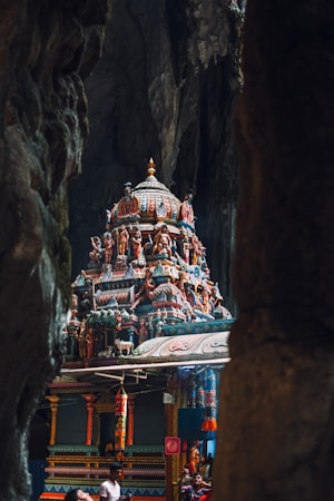 Intricate and colorful Hindu temple carvings are nestled within the dark, dramatic setting of a cave. Statues and artistic embellishments adorn the structure, showcasing traditional architectural designs. Several people are observing and admiring the structure.